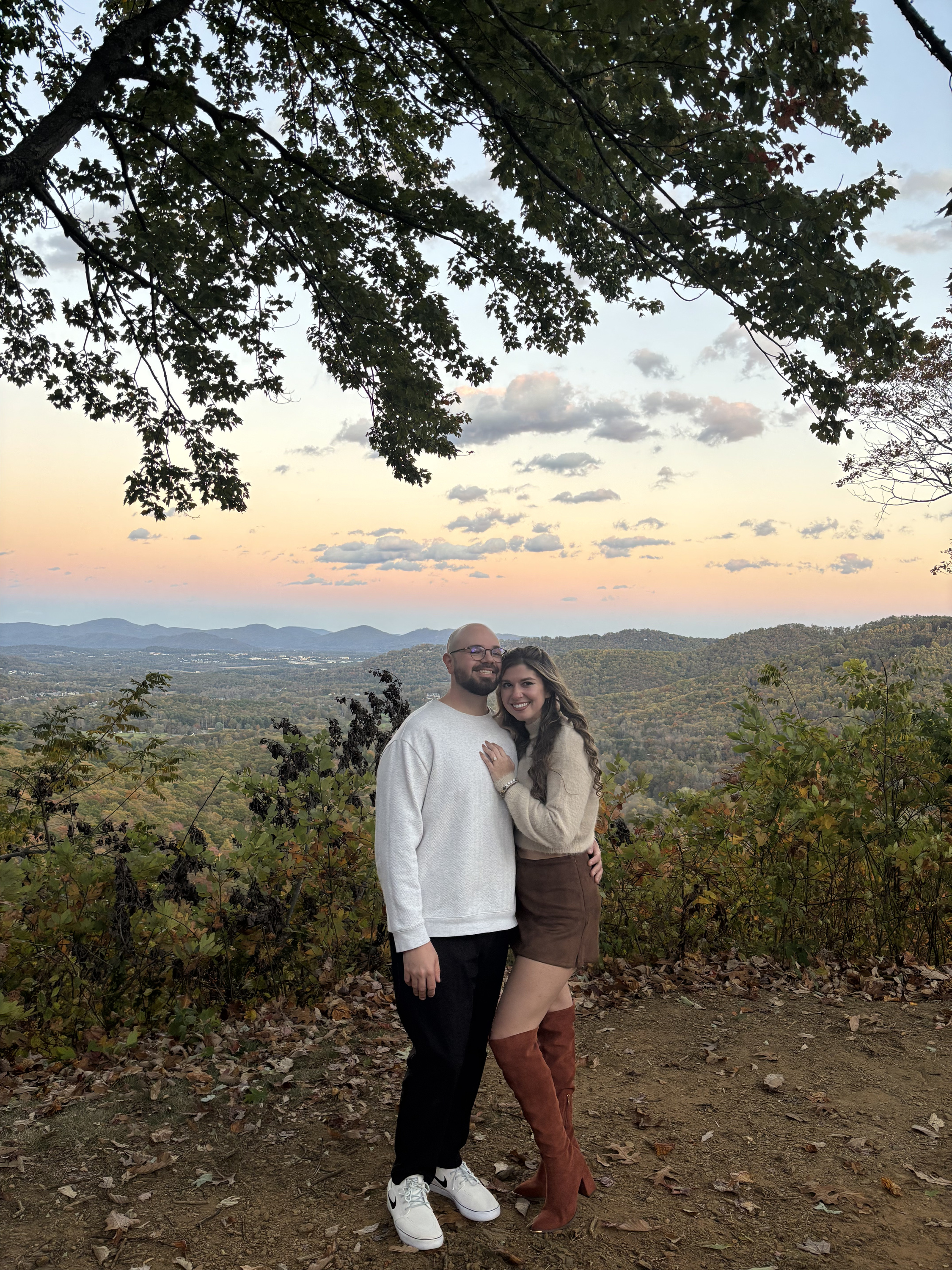 Nicole and Ian at a mountain overlook, arm in arm beneath a leafy branch, with a pale sunset over the ridges behind them.
